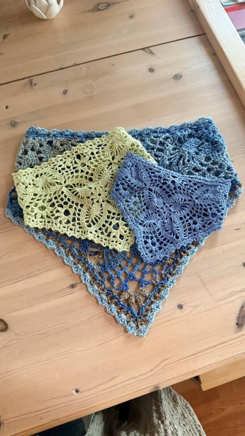 Op de afbeelding: Drie handgemaakte gehaakte sjaals in blauw- en groentinten zijn gerangschikt op een houten oppervlak. De sjaals hebben ingewikkelde open patronen, met een driehoekige vorm. Het kleurenpalet omvat lichtblauw, olijfgroen en donkerdere blauwtinten.