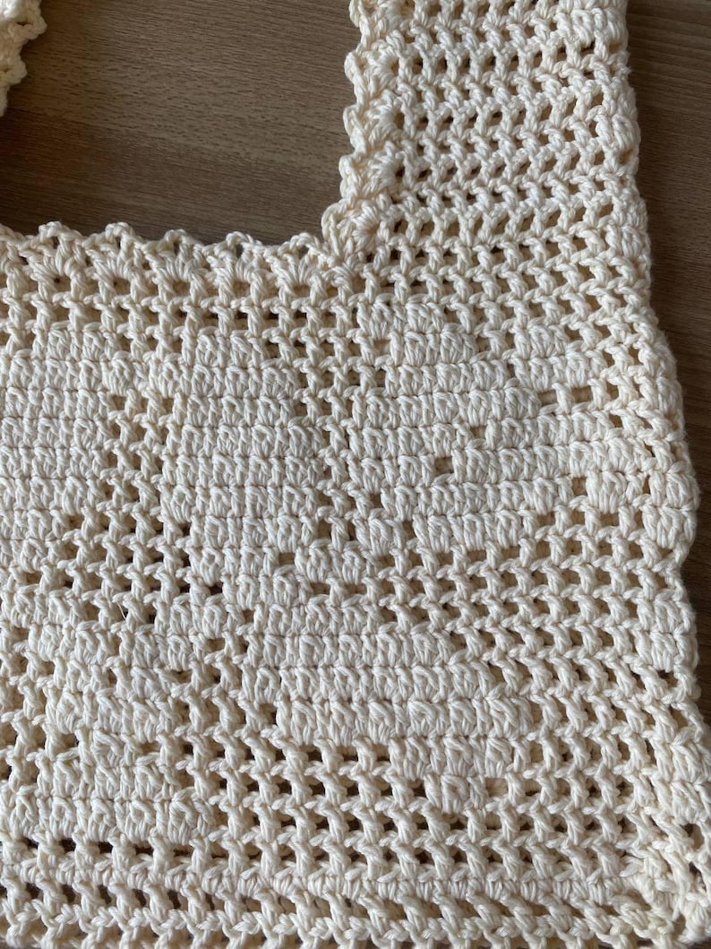 K&ouml;nnte beinhalten: Nahaufnahme eines cremefarbenen H&auml;keltops mit offenem Gewebe. Das Top hat einen gewellten Rand und ein strukturiertes, luftiges Design. Die H&auml;kelarbeit ist detailliert und zeigt das komplizierte Stichmuster und die weiche, nat&uuml;rliche Farbe des Garns.