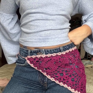 Op de afbeelding: Een diep magenta gehaakte heupsjaal met een geschulpte lichtroze rand. De sjaal wordt gedragen over een blauwe spijkerbroek. De top is een lichtblauw shirt met lange mouwen.