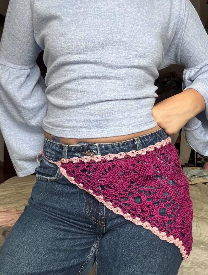 Op de afbeelding: Een diep magenta gehaakte heupsjaal met een geschulpte lichtroze rand. De sjaal wordt gedragen over een blauwe spijkerbroek. De top is een lichtblauw shirt met lange mouwen.