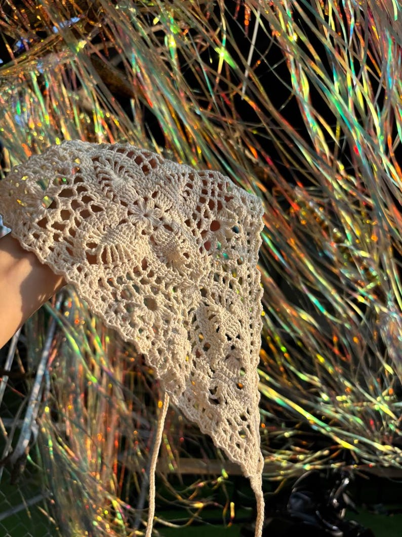 Op de afbeelding: Cr&egrave;mekleurige, handgemaakte gehaakte hoofddoek met een bloemenpatroon. De hoofddoek is driehoekig van vorm met lange banden. Het openwerkontwerp is zichtbaar. De achtergrond is een kleurrijk, iriserend materiaal.