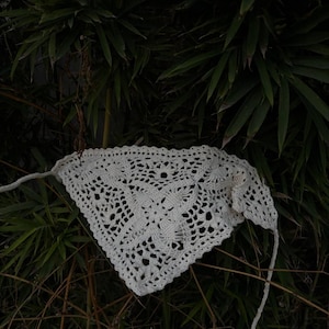 Op de afbeelding: Een witte, gehaakte bandana met een bloemenpatroon. De bandana is driehoekig van vorm en heeft lange banden aan elke kant. De achtergrond is donkergroen met bamboebladeren en -takken.