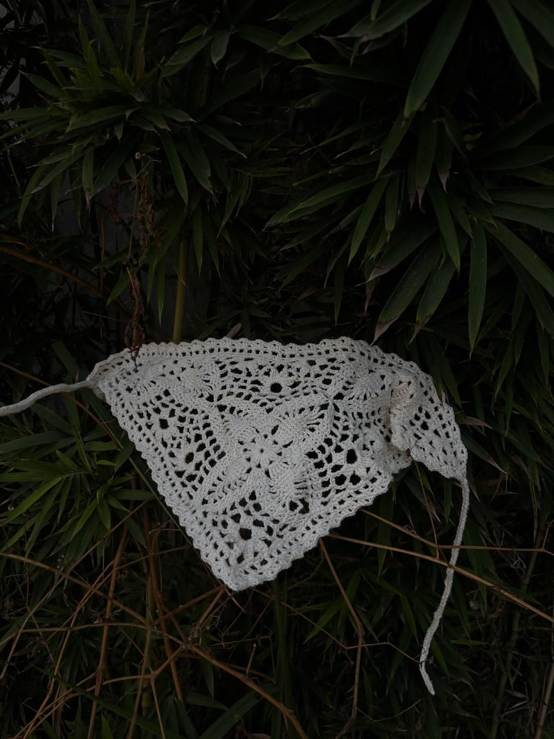 Op de afbeelding: Een witte, gehaakte bandana met een bloemenpatroon. De bandana is driehoekig van vorm en heeft lange banden aan elke kant. De achtergrond is donkergroen met bamboebladeren en -takken.