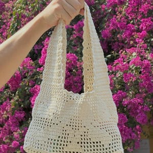 K&ouml;nnte beinhalten: Eine cremefarbene H&auml;keltasche mit offenem Gewebe, rechteckiger Basis und zwei Schultergurten. Die Tasche wird vor einem Hintergrund aus leuchtend rosa Bl&uuml;ten gehalten, was ihre handgefertigte Textur und ihr Design hervorhebt.