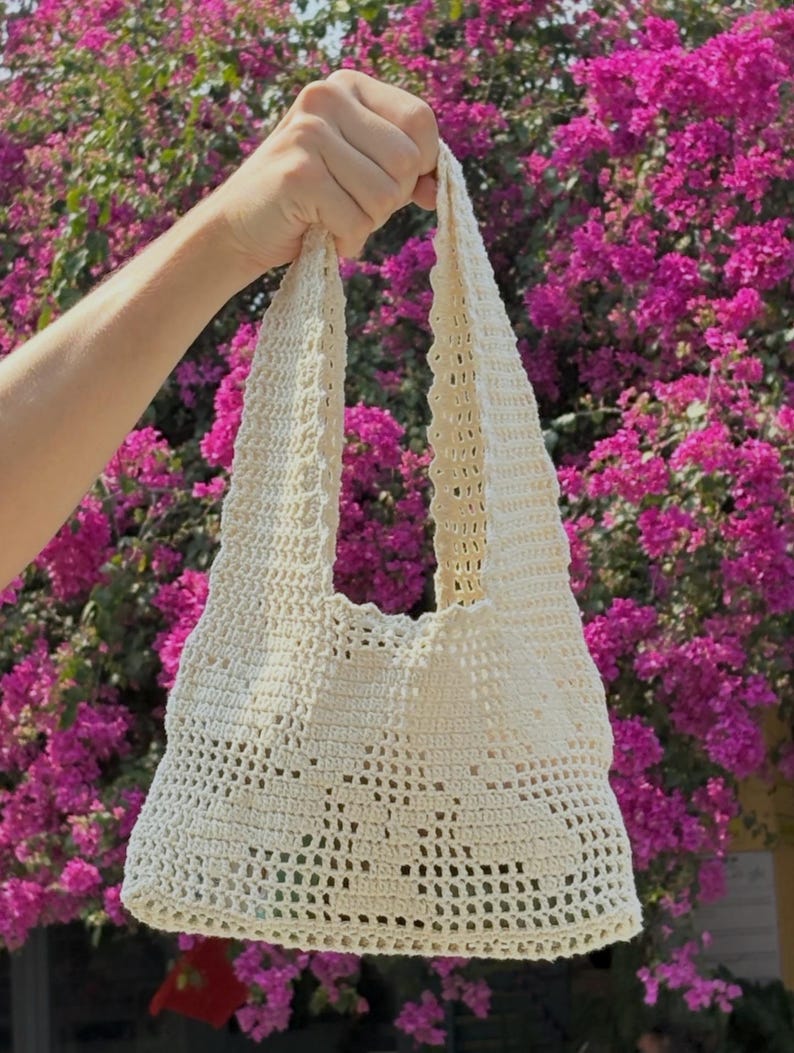 K&ouml;nnte beinhalten: Eine cremefarbene H&auml;keltasche mit offenem Gewebe, rechteckiger Basis und zwei Schultergurten. Die Tasche wird vor einem Hintergrund aus leuchtend rosa Bl&uuml;ten gehalten, was ihre handgefertigte Textur und ihr Design hervorhebt.