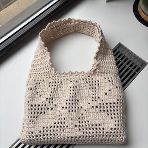 K&ouml;nnte beinhalten: Beige, handgeh&auml;kelte Tragetasche mit einem wellenf&ouml;rmigen oberen Rand. Die Tasche hat ein offenes Design, das ihr ein strukturiertes Aussehen verleiht. Die Tasche ist zum Tragen &uuml;ber der Schulter konzipiert.