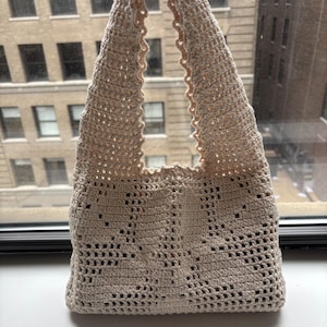 K&ouml;nnte beinhalten: Beige geh&auml;kelte Tragetasche mit dekorativem Lochmuster. Die Tasche hat eine dreieckige Form und einen gewellten Rand. Sie besteht aus weichem, strukturiertem Garn. Geeignet zum Tragen verschiedener Gegenst&auml;nde.