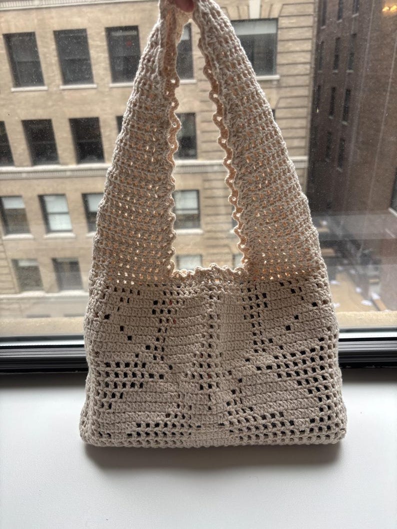 K&ouml;nnte beinhalten: Beige geh&auml;kelte Tragetasche mit dekorativem Lochmuster. Die Tasche hat eine dreieckige Form und einen gewellten Rand. Sie besteht aus weichem, strukturiertem Garn. Geeignet zum Tragen verschiedener Gegenst&auml;nde.