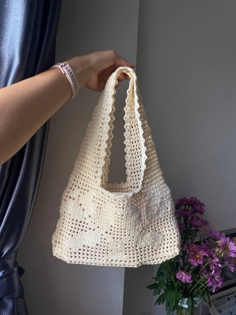 K&ouml;nnte beinhalten: Cremefarbene H&auml;keltasche mit gewelltem Rand. Die Tasche hat einen rechteckigen K&ouml;rper und zwei Henkel. Die Tasche wird von einer Hand gehalten. Im Hintergrund befindet sich ein Strau&szlig; lila Blumen.