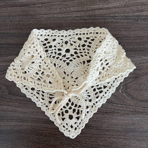 Op de afbeelding: Cr&egrave;mekleurige, gehaakte halsdoek met een vierkante vorm en een ingewikkeld opengewerkt ontwerp. De sjaal heeft een striksluiting aan de voorkant en is gemaakt van zacht, getextureerd garen. Het item is waarschijnlijk handgemaakt.