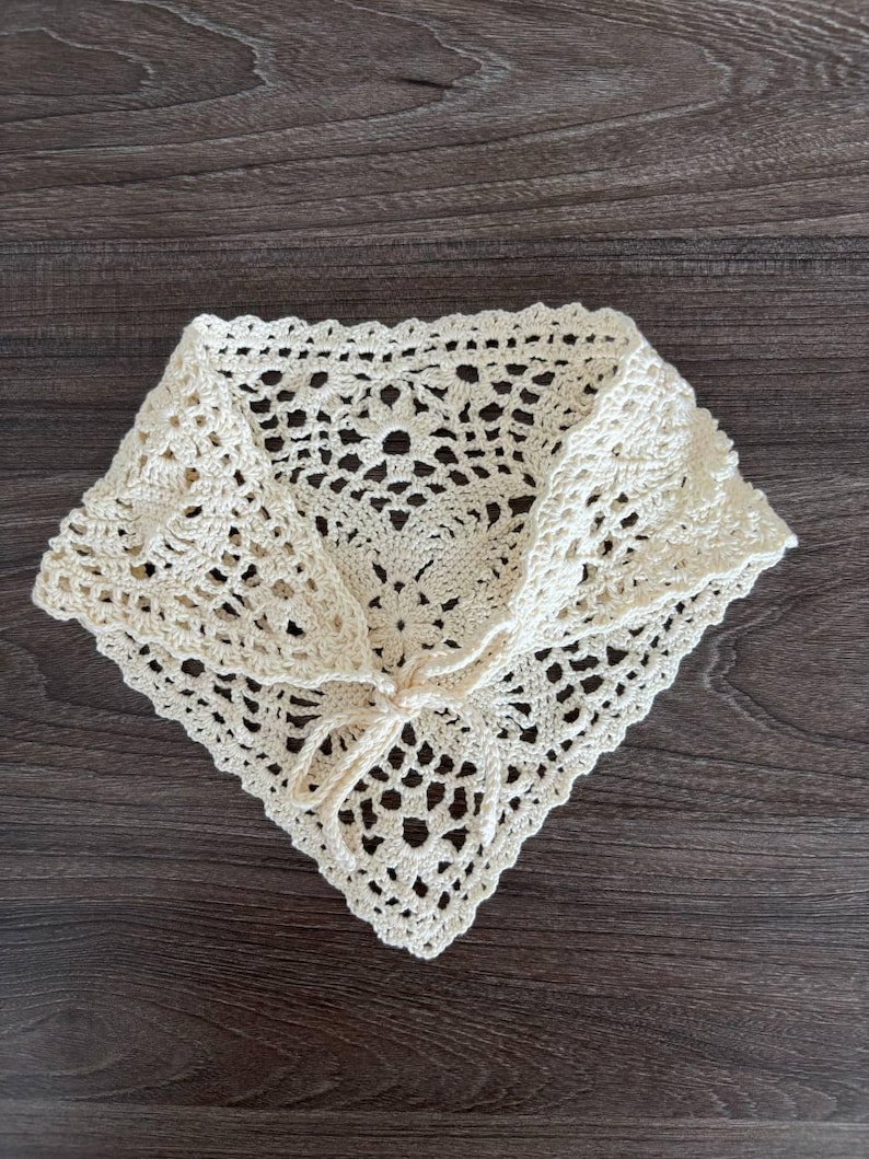 Op de afbeelding: Cr&egrave;mekleurige, gehaakte halsdoek met een vierkante vorm en een ingewikkeld opengewerkt ontwerp. De sjaal heeft een striksluiting aan de voorkant en is gemaakt van zacht, getextureerd garen. Het item is waarschijnlijk handgemaakt.
