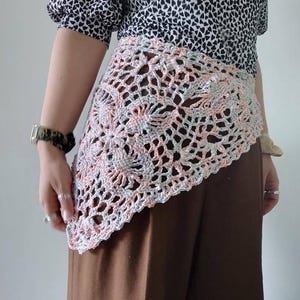 Op de afbeelding: Een gehaakte omslagdoek in pastelkleuren roze, grijs en wit. De omslagdoek heeft een bloemenpatroon en wordt gedragen over een bruine broek en een zwart-wit patroon top. Een uniek modeaccessoire.