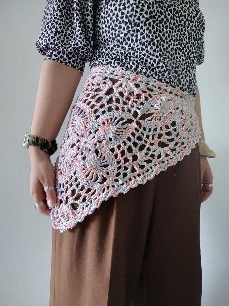Op de afbeelding: Een gehaakte omslagdoek in pastelkleuren roze, grijs en wit. De omslagdoek heeft een bloemenpatroon en wordt gedragen over een bruine broek en een zwart-wit patroon top. Een uniek modeaccessoire.