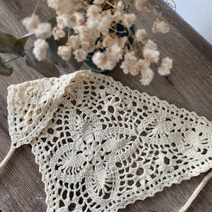 Op de afbeelding: Cr&egrave;mekleurige, handgemaakte gehaakte hoofddoek met een bloemmotief. De hoofdbedekking is driehoekig, met een delicaat openwerkontwerp en lange banden. De sjaal wordt gepresenteerd op een houten oppervlak, met een vaas met gedroogde bloemen op de achtergrond.