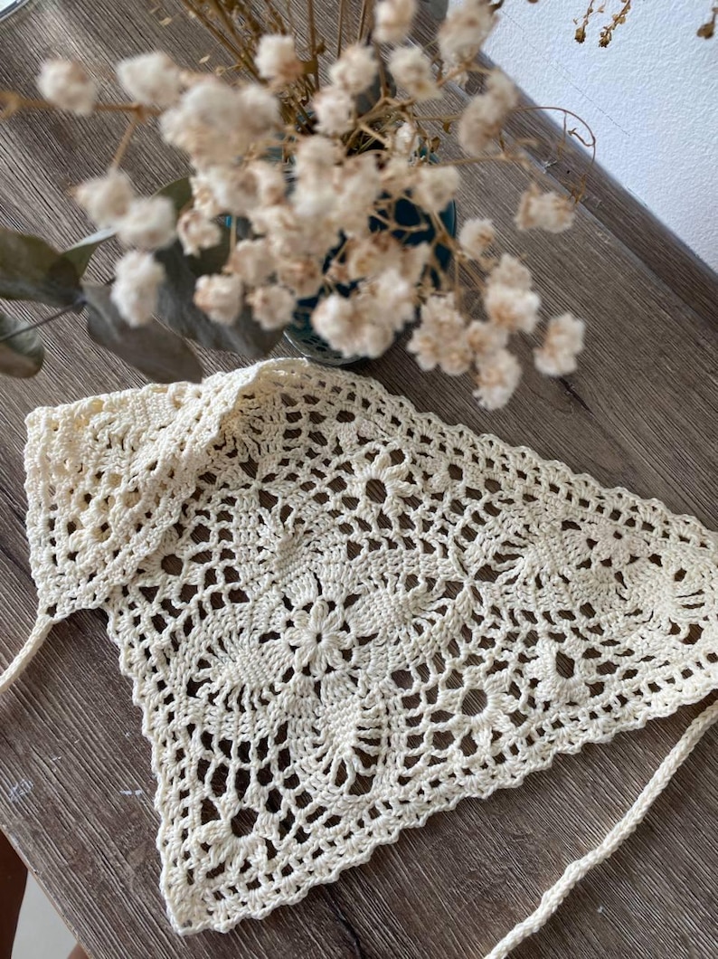 Op de afbeelding: Cr&egrave;mekleurige, handgemaakte gehaakte hoofddoek met een bloemmotief. De hoofdbedekking is driehoekig, met een delicaat openwerkontwerp en lange banden. De sjaal wordt gepresenteerd op een houten oppervlak, met een vaas met gedroogde bloemen op de achtergrond.