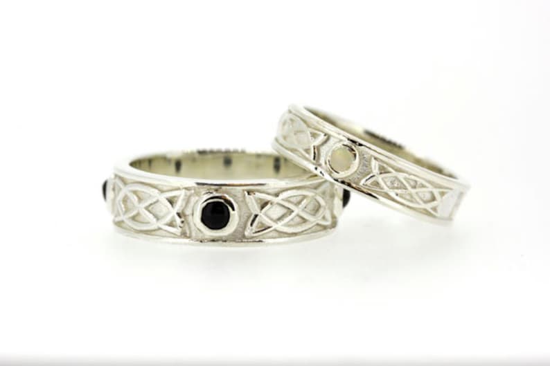 May include: Two silver Celtic knot wedding bands. One band features black gemstones and the other has a white gemstone.