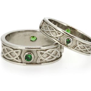 May include: Two silver Celtic knot wedding bands with green gemstones. The bands are intertwined and feature a detailed knot design.
