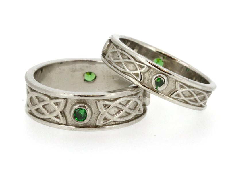 May include: Two silver Celtic knot wedding bands with green gemstones. The bands are intertwined and feature a detailed knot design.