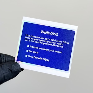 以下が含まれることがあります： 青い背景に「WINDOWS」の文字が上部に書かれたステッカー。ステッカーには「お使いのコンピュータで致命的なエラーが発生しました。これは、オペレーティングシステムがひどいからです。」と書かれています。
