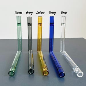 Puede incluir: Cinco pajitas de vidrio en verde, gris, ámbar, azul marino y transparente. Cada pajita tiene una punta doblada y un cuerpo recto. Los nombres de los colores están impresos encima de cada pajita.