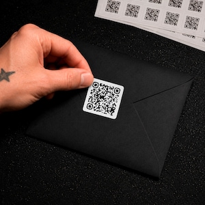 May include: A square white sticker featuring a black QR code pattern being applied to a black envelope. A sheet of identical white QR code stickers is visible in the upper portion of the frame, showcasing multiple individual square labels.