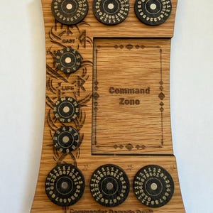 Könnte beinhalten: Ein Spiel-Tracker-Board aus Holz mit einem rechteckigen Command Zone-Bereich und mehreren kreisförmigen Drehreglern. Das Board enthält eingravierte Textbeschriftungen wie Commander Damage Received, Cast, Life und Commander Damage Dealt.