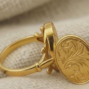 May include: A gold ring with an oval, hinged locket. The locket and ring band feature intricate detailing. The locket is open, revealing an engraved floral design. The ring is set against a neutral fabric background.