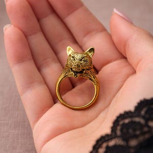 May include: A gold-coloured ring with a detailed wolf head design. The ring's head has intricate detailing, including the wolf's ears, eyes, and open mouth. The ring is held in a hand, showcasing its design and craftsmanship.