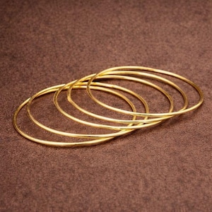 Könnte beinhalten: Fünf dünne, goldfarbene Armreifen werden gezeigt. Die kreisförmigen Armreifen, wahrscheinlich aus Metall, sind auf einer braunen Oberfläche angeordnet. Das Licht reflektiert sich auf der glatten Oberfläche der Armreifen und erzeugt einen einfachen und eleganten Look. Jeder Armreif hat einen Durchmesser von etwa 20 cm.