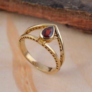 May include: A gold ring featuring a teardrop-shaped, deep red gemstone. The ring has a double-band design with small, evenly spaced holes along the outer band. The gemstone is set in a gold bezel.