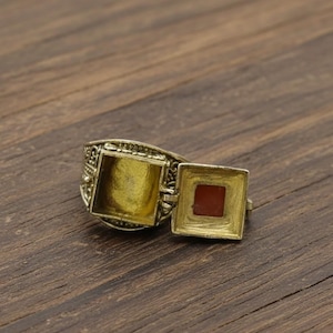Puede incluir: Un anillo antiguo de color dorado con un compartimento cuadrado abierto y un compartimento cuadrado más pequeño con una piedra roja. El anillo tiene detalles intrincados alrededor de la banda y los compartimentos. El anillo se muestra sobre una superficie de madera.