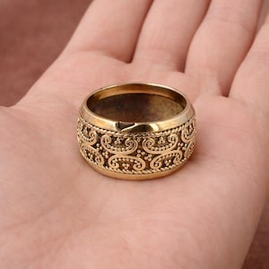 May include: A gold-colored ring with an ornate, repeating pattern of swirls and dots. The ring is thick and has a slightly worn appearance, suggesting it may be vintage or antique. The ring is shown on a person's palm.