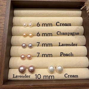 May include: A display of pearl stud earrings in a wooden tray. The earrings are in various colors, including cream, champagne, lavender, and peach. Sizes range from 6 mm to 10 mm. The text on the tray indicates the color and size of each pair.