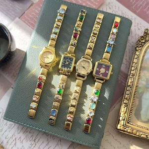 Może przedstawiać: Cztery złote zegarki na rękę z ozdobnymi paskami i różnymi tarczami, w tym okrągłymi, kwadratowymi i owalnymi. Paski mają kolorowe zdobienia. Zegarki są ułożone na zielonej powierzchni.