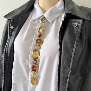 May include: A distinctive necklace composed of assorted vintage watch faces in gold and burgundy hues. The watch faces are linked vertically, forming a striking accessory. The necklace is displayed over a white collared shirt and a black leather jacket.
