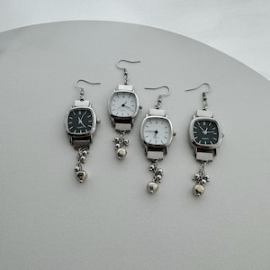 May include: Four earrings designed as miniature wristwatches with silver-toned metal bands and square faces. Two watches have black dials and two have white dials. Each earring features a dangling chain with small silver-toned metal beads.