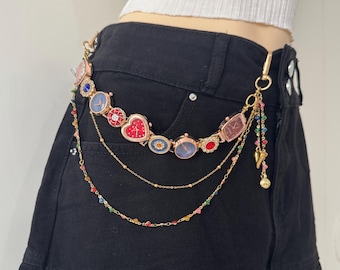 May include: A gold-colored chain belt with multiple watch-shaped charms in various colors and designs. The belt features a heart-shaped charm and a dangling charm with colorful beads and a gold-colored pendant. The belt is attached to black shorts.