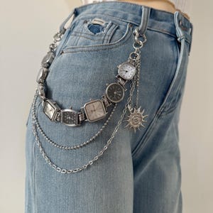May include: Silver watch chain belt with multiple square and rectangular watch faces. The belt is attached to light blue denim jeans. A sun charm and dangling chains add detail to the accessory.