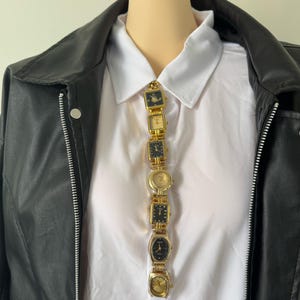 Peut inclure: Un collier doré composé de plusieurs montres vintage avec différentes formes et designs de cadrans. Les montres sont reliées verticalement, portées sur une chemise blanche à col et une veste en cuir noire.