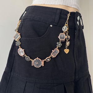 Puede incluir: Una cadena de reloj dorada, adornada con pequeñas esferas y dijes decorativos, está sujeta a la cintura de unos pantalones cortos negros. La cadena presenta varios diseños de relojes, incluyendo esferas redondas, cuadradas y rectangulares, además de dijes florales y en forma de corazón.