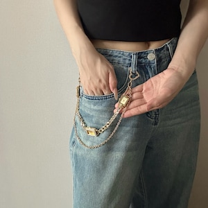 May include: Gold-toned watch chain with two rectangular watch faces and a heart-shaped clasp. The chain is attached to a pair of light blue denim jeans. The watch faces have a square shape.