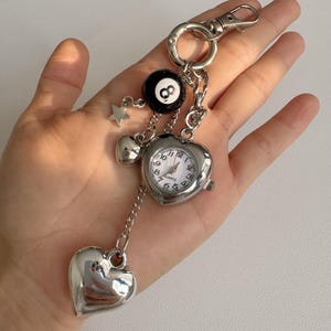 Könnte beinhalten: Ein silberfarbener Schlüsselanhänger mit einer herzförmigen analogen Quarzuhr mit weißem Zifferblatt, einem schwarzen Acht-Ball-Anhänger, einem kleinen silbernen Stern und zwei glatten silbernen Herzanhängern an einem runden Ring mit Karabiner.