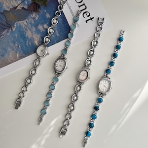 May include: Four silver-toned bracelet watches with oval faces and white dials. Each watch features a different gemstone or pearl accent along the bracelet. The watches are displayed on a white surface.