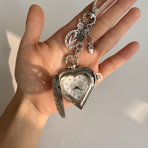May include: A silver heart-shaped pocket watch with a quartz movement, attached to a keychain. The watch face displays black numerals on a white background. The watch is open, revealing the time. Decorative charms dangle from the keychain.