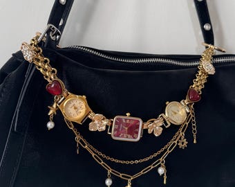 May include: Black handbag adorned with a gold chain featuring three vintage watches with red and gold details. The chain includes heart charms, floral elements, and dangling charms such as pearls and a key.