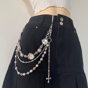May include: Silver-toned belt chain with multiple watch faces, pearl accents, and a cross pendant. The chain is attached to a black skirt. The watches have white faces and are surrounded by decorative metalwork.
