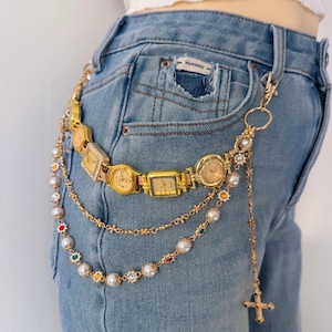 Puede incluir: Una cadena de cinturón dorada con múltiples esferas de reloj vintage, perlas y detalles decorativos. La cadena está sujeta a unos vaqueros azules. Un colgante de cruz cuelga de la cadena.