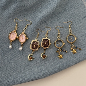 May include: Three pairs of gold-tone watch earrings with fishhook wires. The first pair has oval pink faces and white pearl drops. The second pair has rectangular brown faces and crescent moon charms. The third pair has round blue faces and gold star charms.