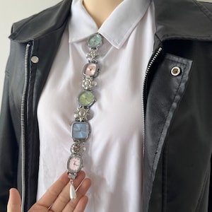 May include: A unique necklace featuring a series of small, colorful square-faced watches in pastel shades of pink, blue, and green. The watches are connected by a silver chain with decorative elements and a dangling pearl. The necklace is worn over a white collared shirt and black leather jacket.