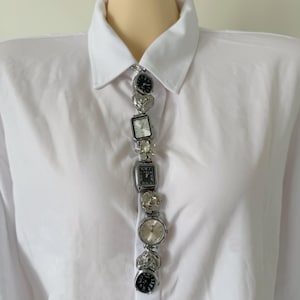 Peut inclure: Une chemise blanche à col avec un broche montre argentée unique. La broche est composée de plusieurs petites montres de style vintage de différentes formes et designs, reliées verticalement.